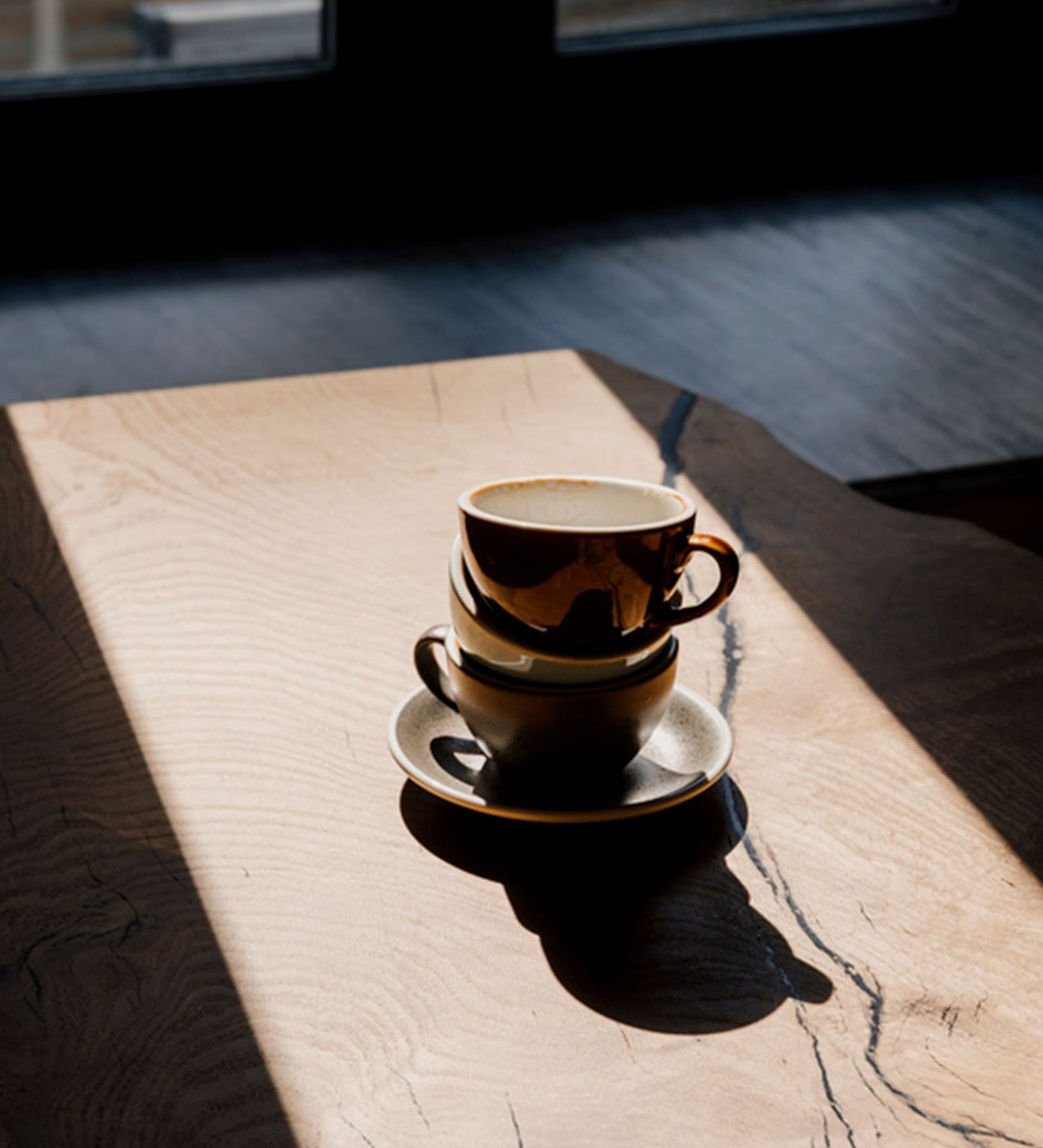 A carved wood coffee table with a 3 coffee used coffee cups stacked upon a plate lit by natural sunlight