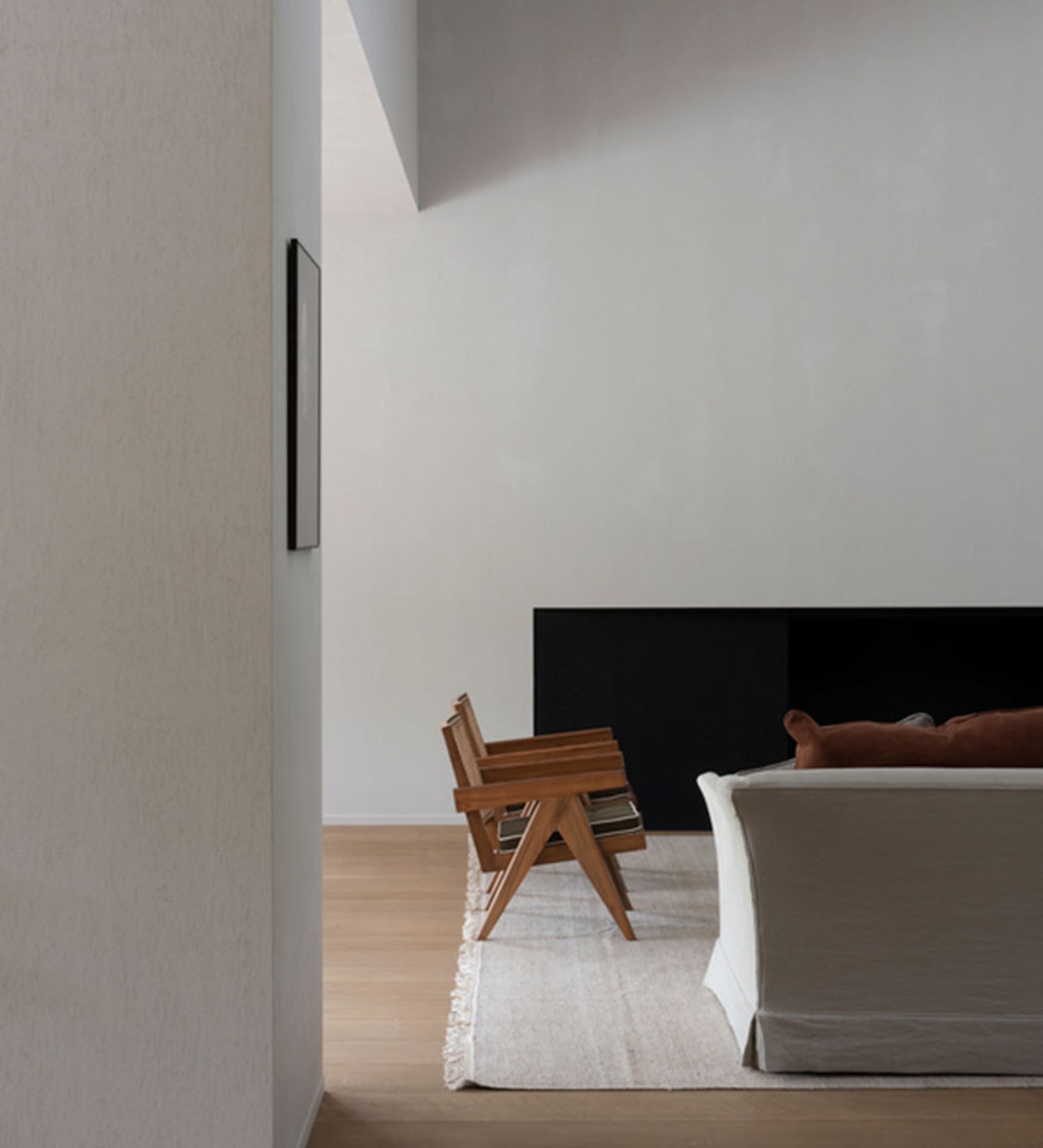 Half view of a sitting area with white calls, wood flooring, white rug, the back of a white covered sofa, and 2 wood chairs placed perpendicular to the sofa