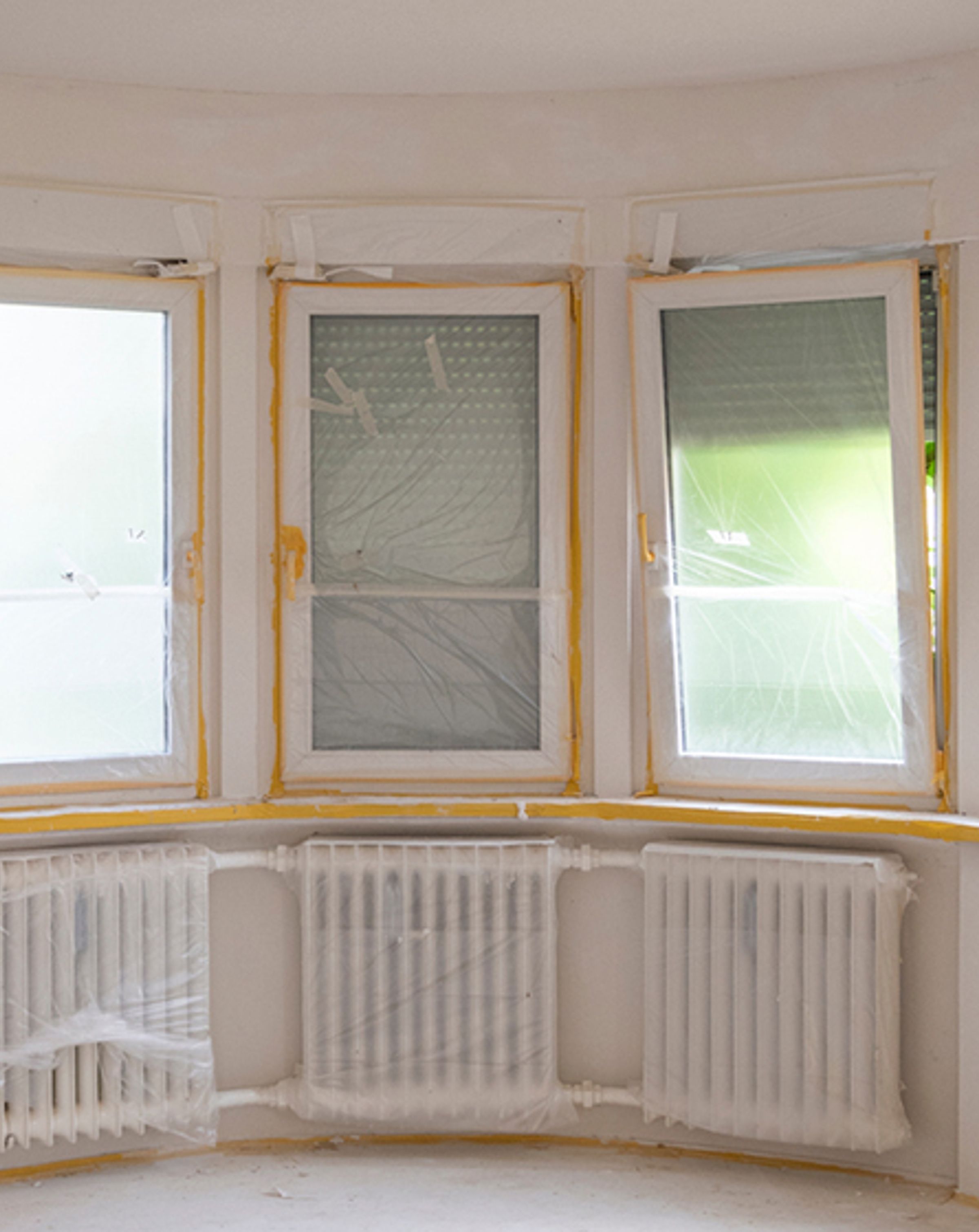 Three windows with orange masking tap around around them, with a bar heater under each covered in plastic, in preparation for the room to be painted