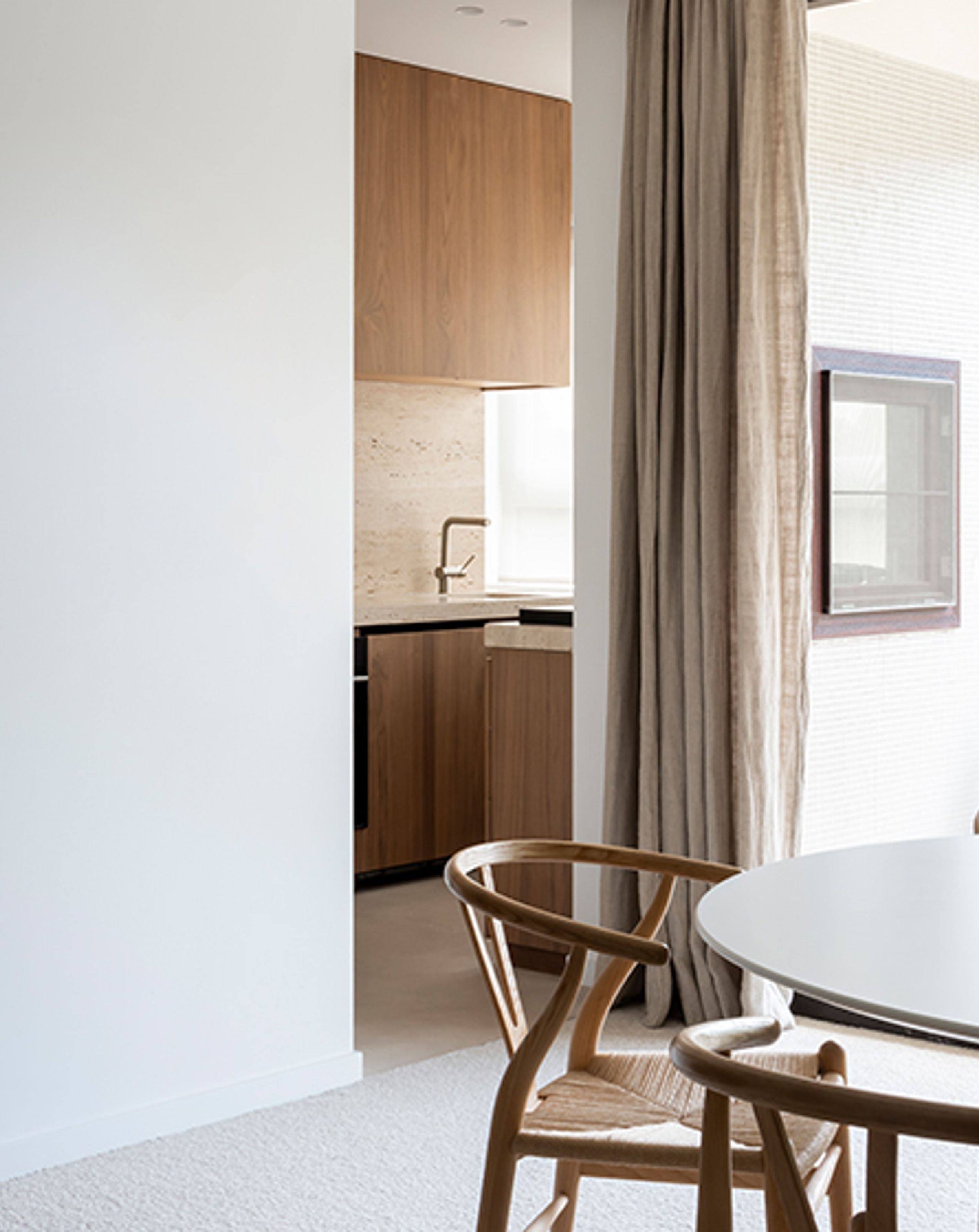 White table with wooden chairs in the foreground, a white wall behind, and an opening to a kitchen with wooden cabinetry and a brass coloured tap