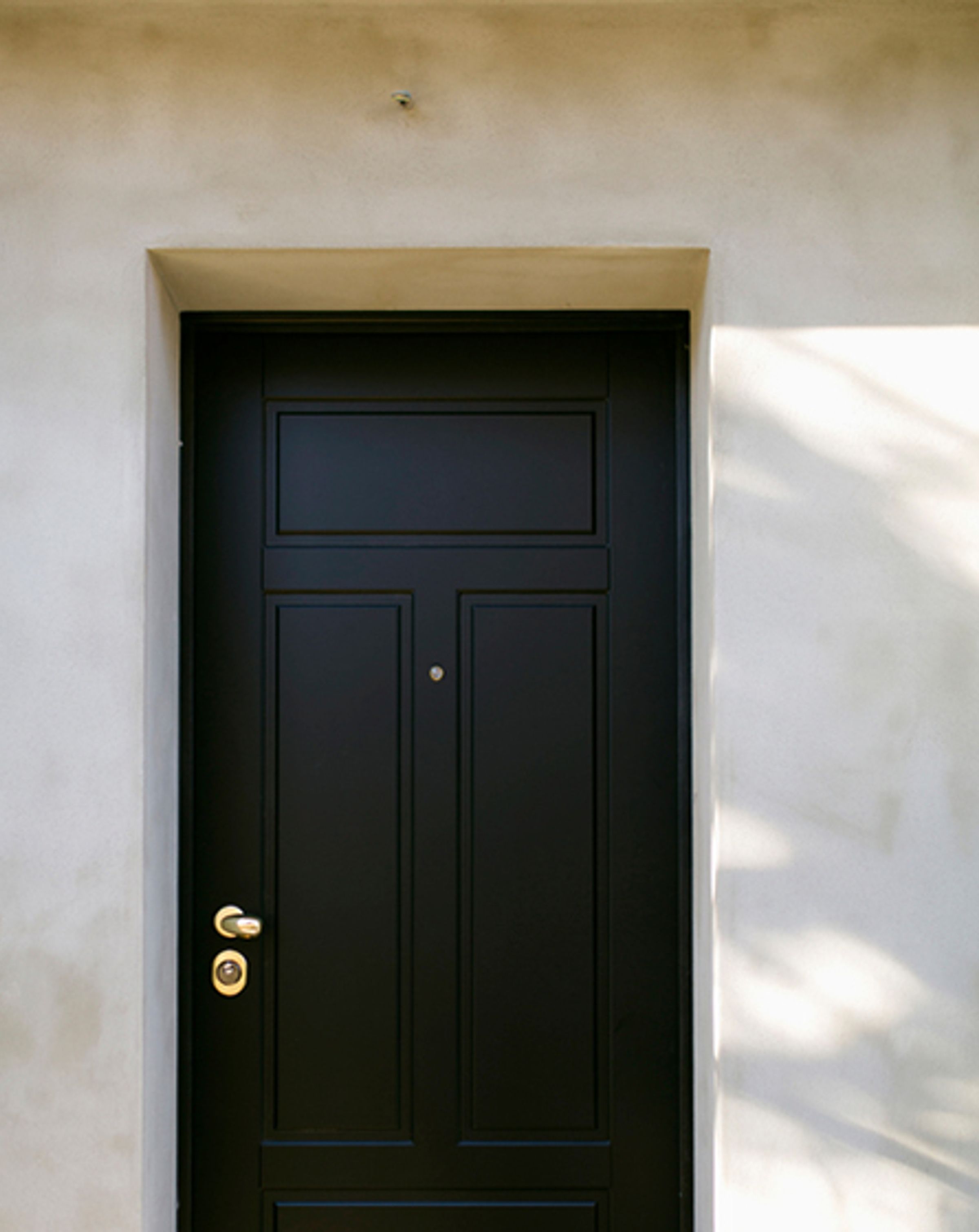 A white stone wall with a large black wooden door, with bronze handle on the left side and keyhole below