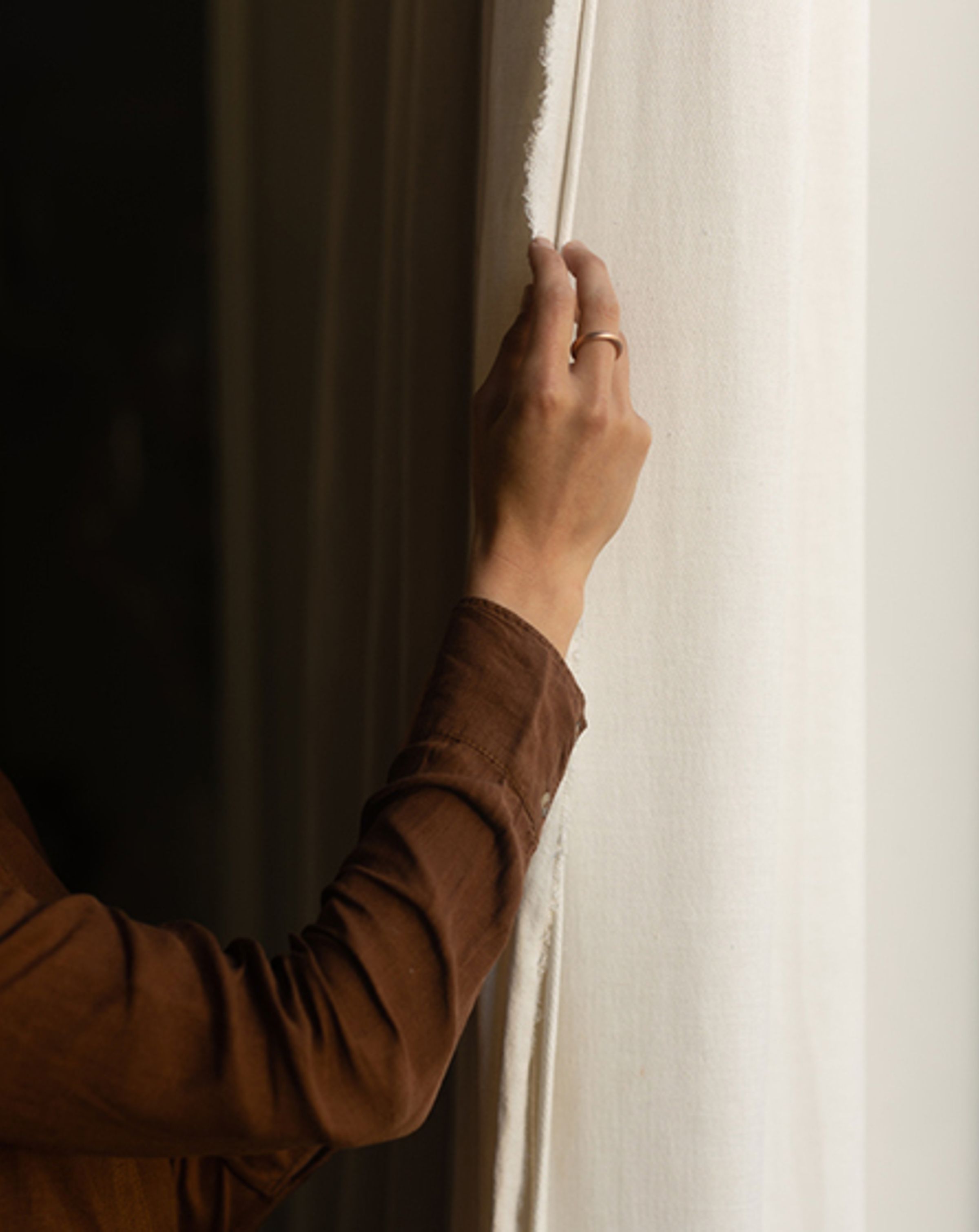 A person pulling back a curtain using their right hand, wearing a dark brown long sleeve shirt.